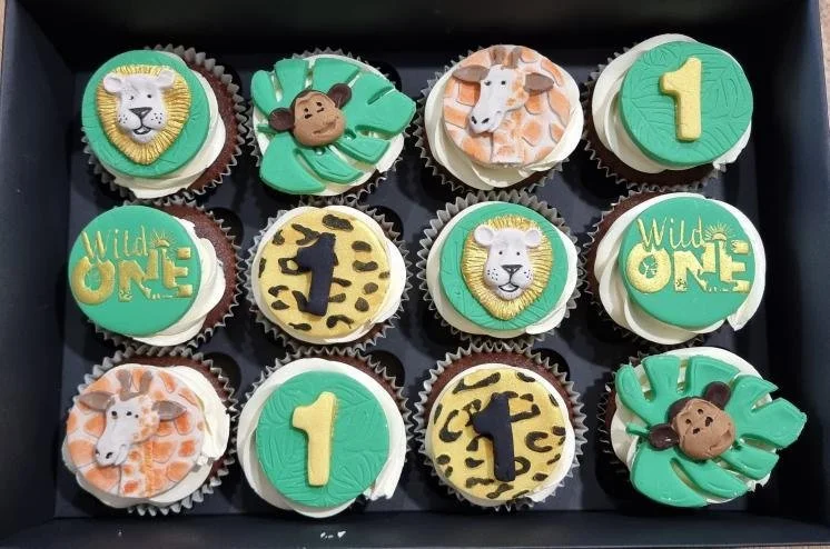 A box of twelve cupcakes with jungle animal themed decorations and the number one, for a first birthday celebration.