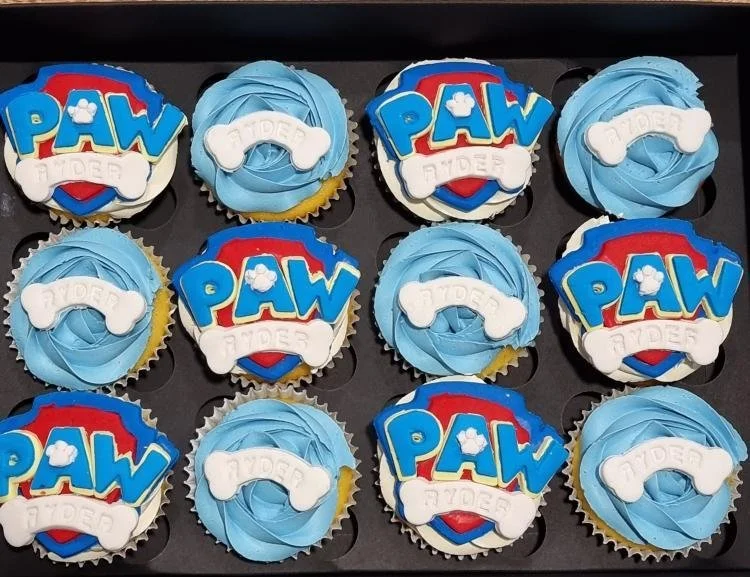 A tray of 12 cupcakes decorated with Paw Patrol theme, featuring blue swirl frosting and Paw Patrol logo toppers with the words 'PAW' and a bone.