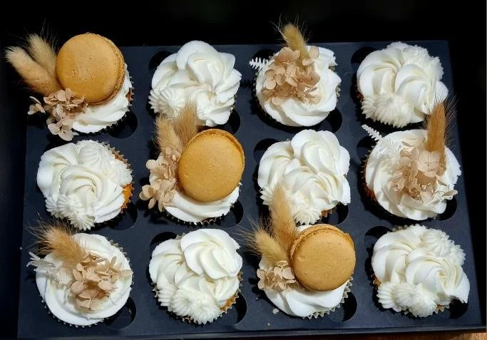 A black tray holding twelve decorated cupcakes with white frosting, beige floral decorations, and macarons on top.