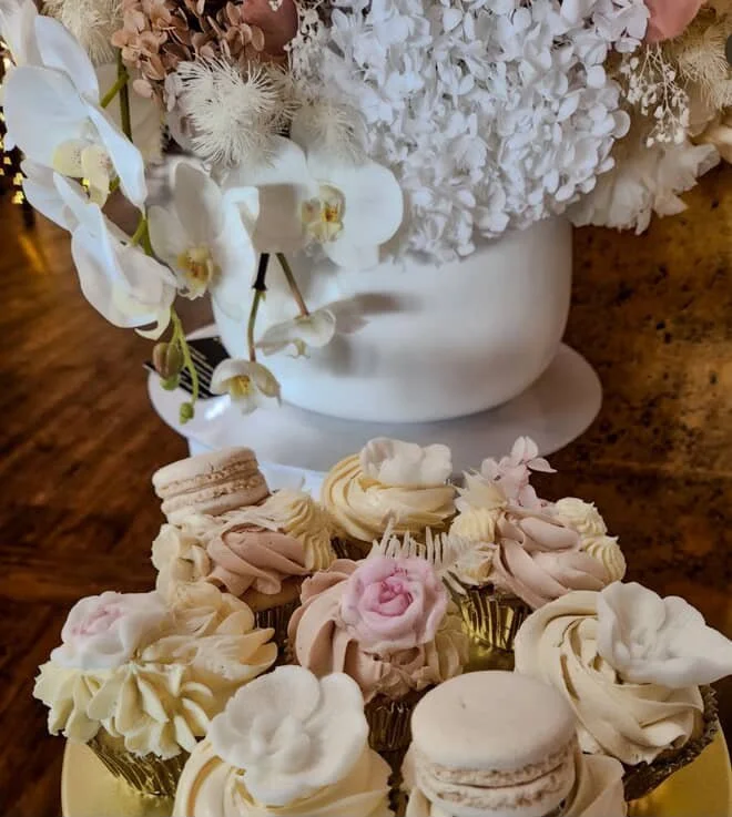 Assorted decorated cupcakes with floral decorations and macarons on a gold tray, between a white floral arrangement in a white vase on a wooden surface.