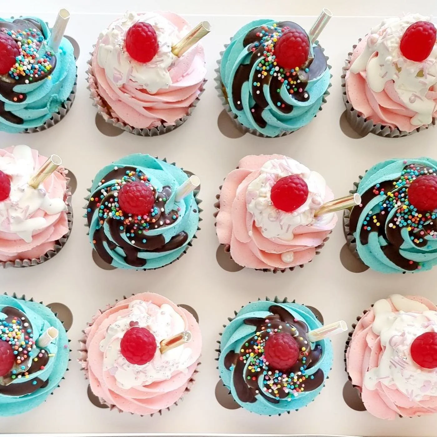 Arrangement of colorful cupcakes with pink, blue, and white frosting, decorated with sprinkles, cherries, and white chocolate sticks.