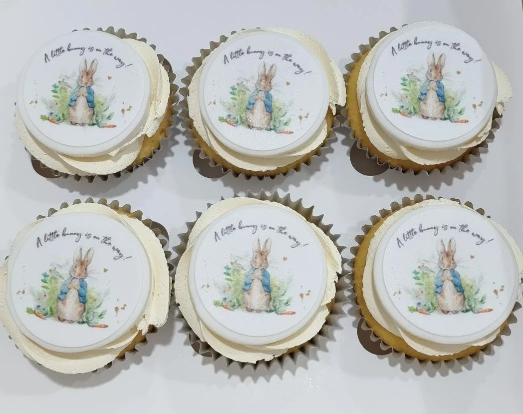 Six cupcakes with white frosting featuring a printed image of a rabbit wearing a blue jacket, surrounded by greenery and small hearts, with the phrase "A little bunny is on the way!" written on top.