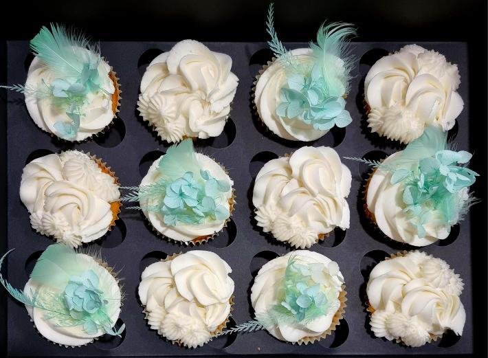 A tray of twelve cupcakes decorated with white frosting and light blue flower and feather decorations.