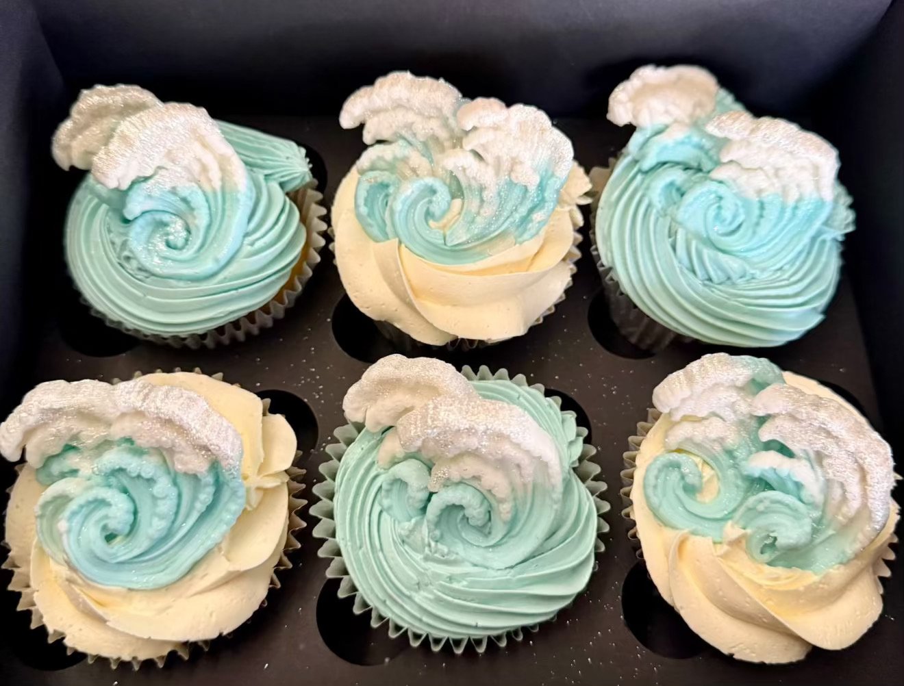 Six cupcakes with aqua-colored swirled frosting and white powdered sugar on top, arranged in a black box.