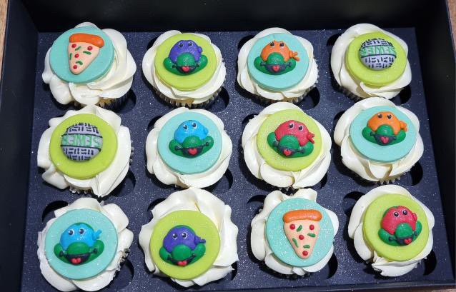 Twelve cupcakes with colorful alien, watermelon, and pizza-themed toppers in a box.