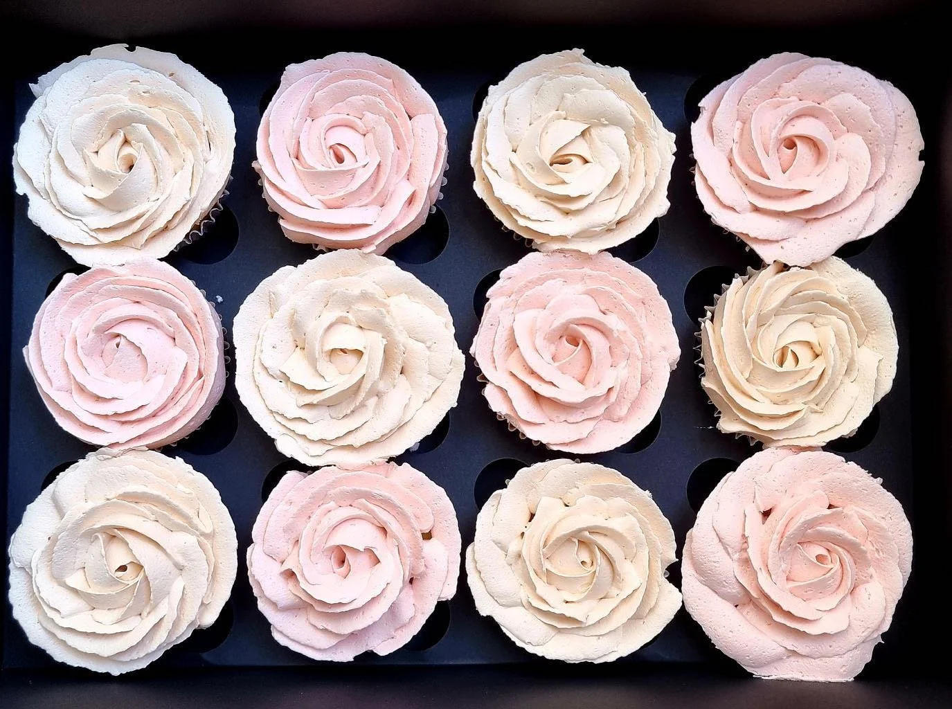 Tray of 12 cupcakes decorated with pink and white frosting in rose shapes.