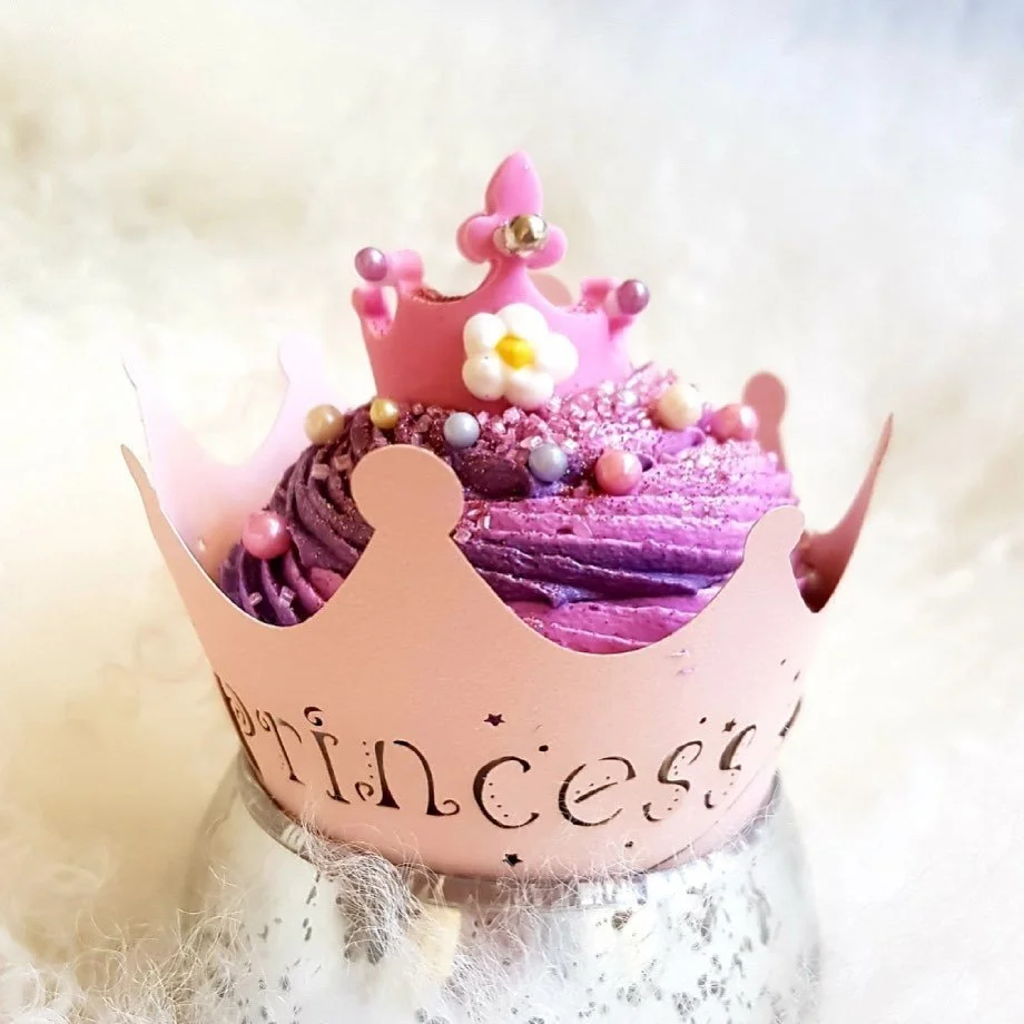 Pink and purple cupcake decorated with a pink crown, a small white flower, and colorful sprinkles, wrapped in a pink crown-shaped paper with 'princess' printed on it, placed on a soft, fuzzy surface.