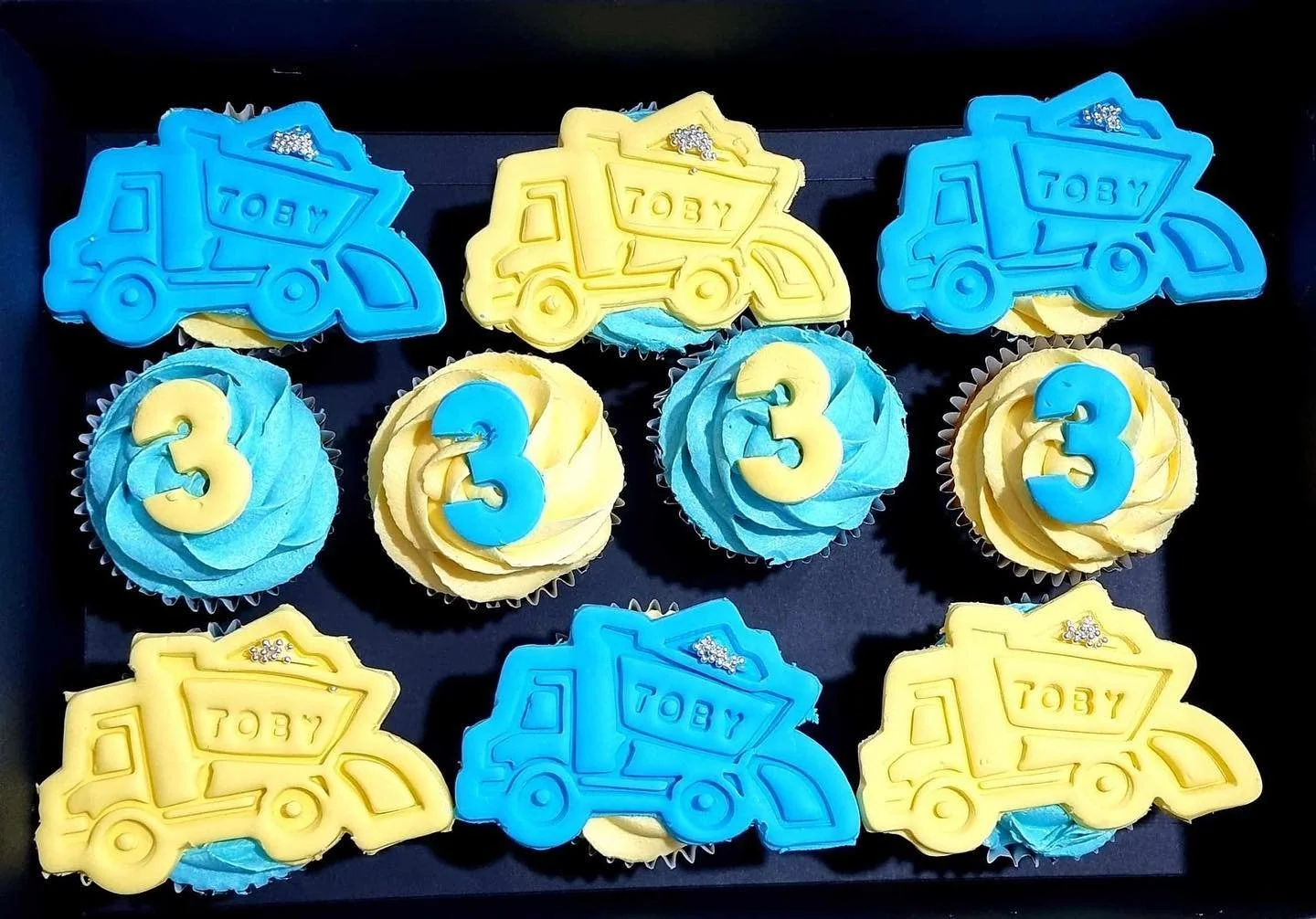 Cupcakes decorated with blue and yellow frosting, featuring a number three and construction truck toppers that say by.