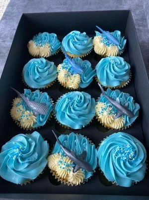 Box of twelve cupcakes decorated with blue frosting, sprinkles, and shark-shaped toppers for a themed party.