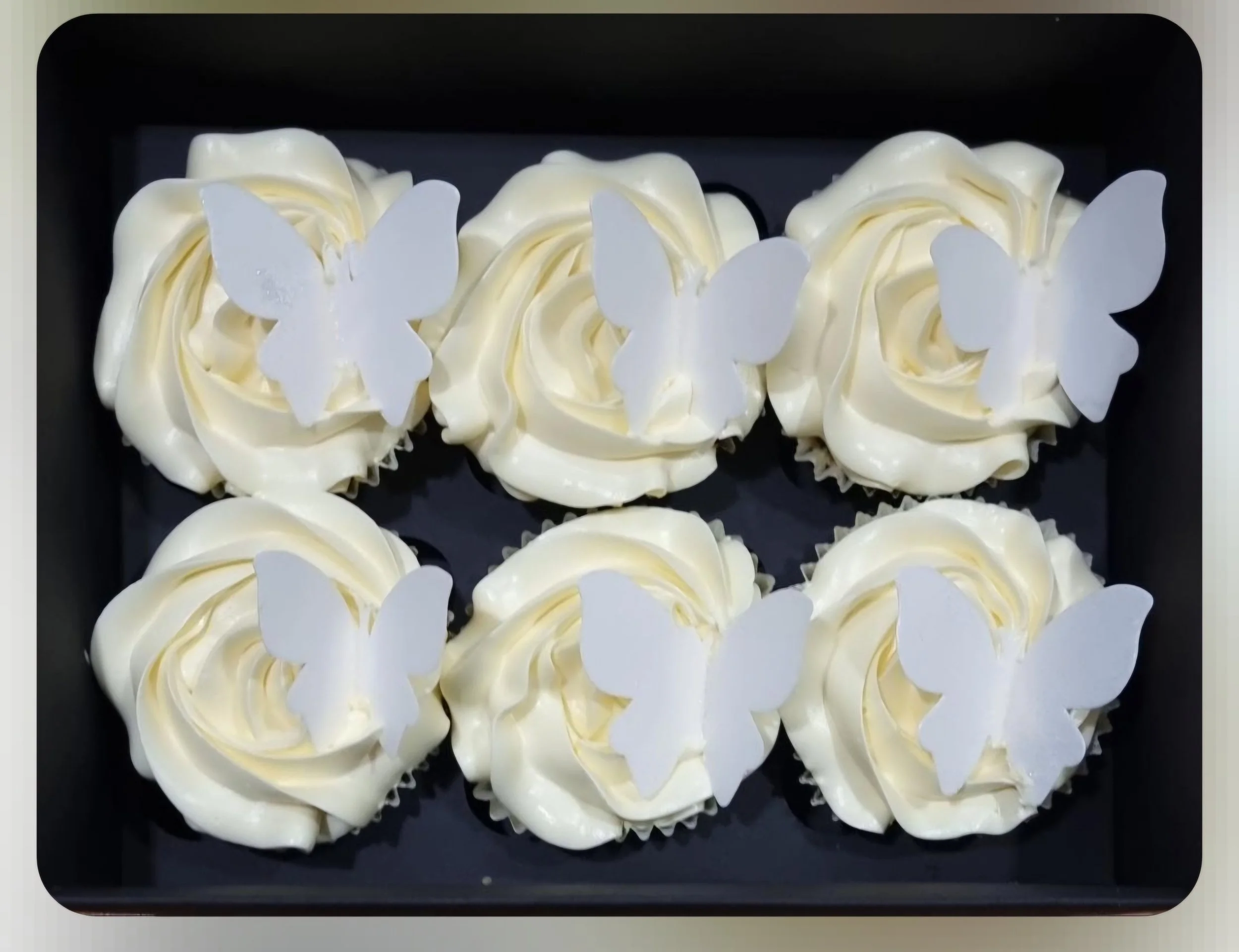 Multiple cream-colored cupcakes with white butterfly toppers, arranged on a black tray.