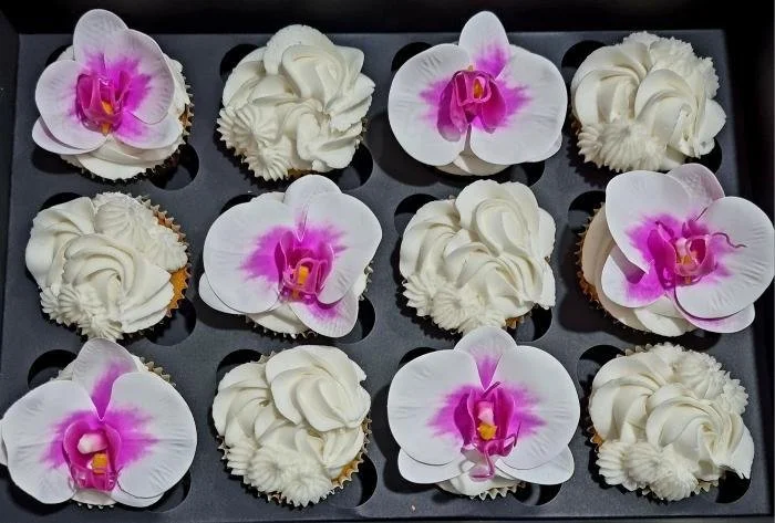A tray of twelve cupcakes decorated with white frosting and topped with pink and white artificial orchids.