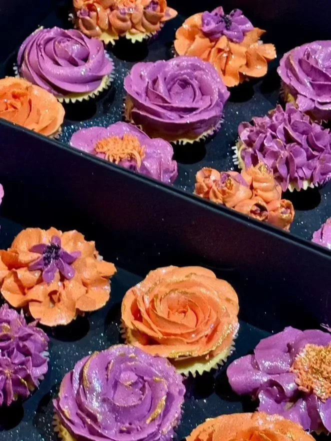 Cupcakes decorated with orange and purple frosting styled like roses and flowers, with some glitter accents, arranged in a black box.