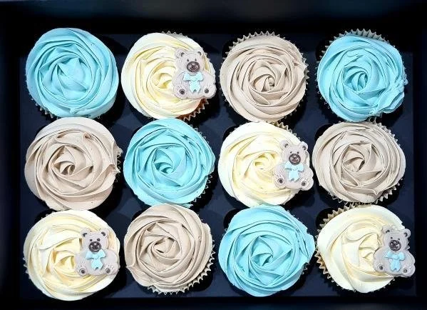 Box of 12 cupcakes decorated with swirl frosting in blue, beige, and yellow, some with bear and honey jar toppers.