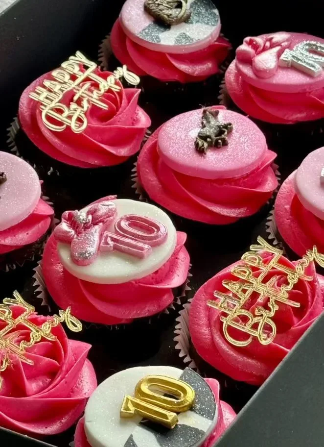A box of pink cupcakes decorated for a 10th birthday celebration, each with unique toppers including number 10, 'Happy Birthday' message, and various figurines and decorations.