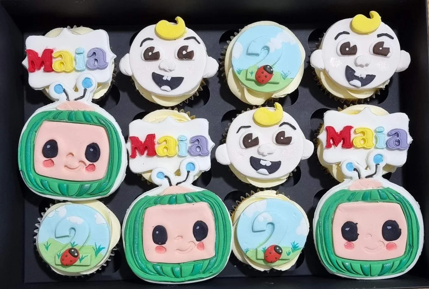 Decorated cupcakes with cartoon character faces, ladybug, cloud, and number 2, in a black box.