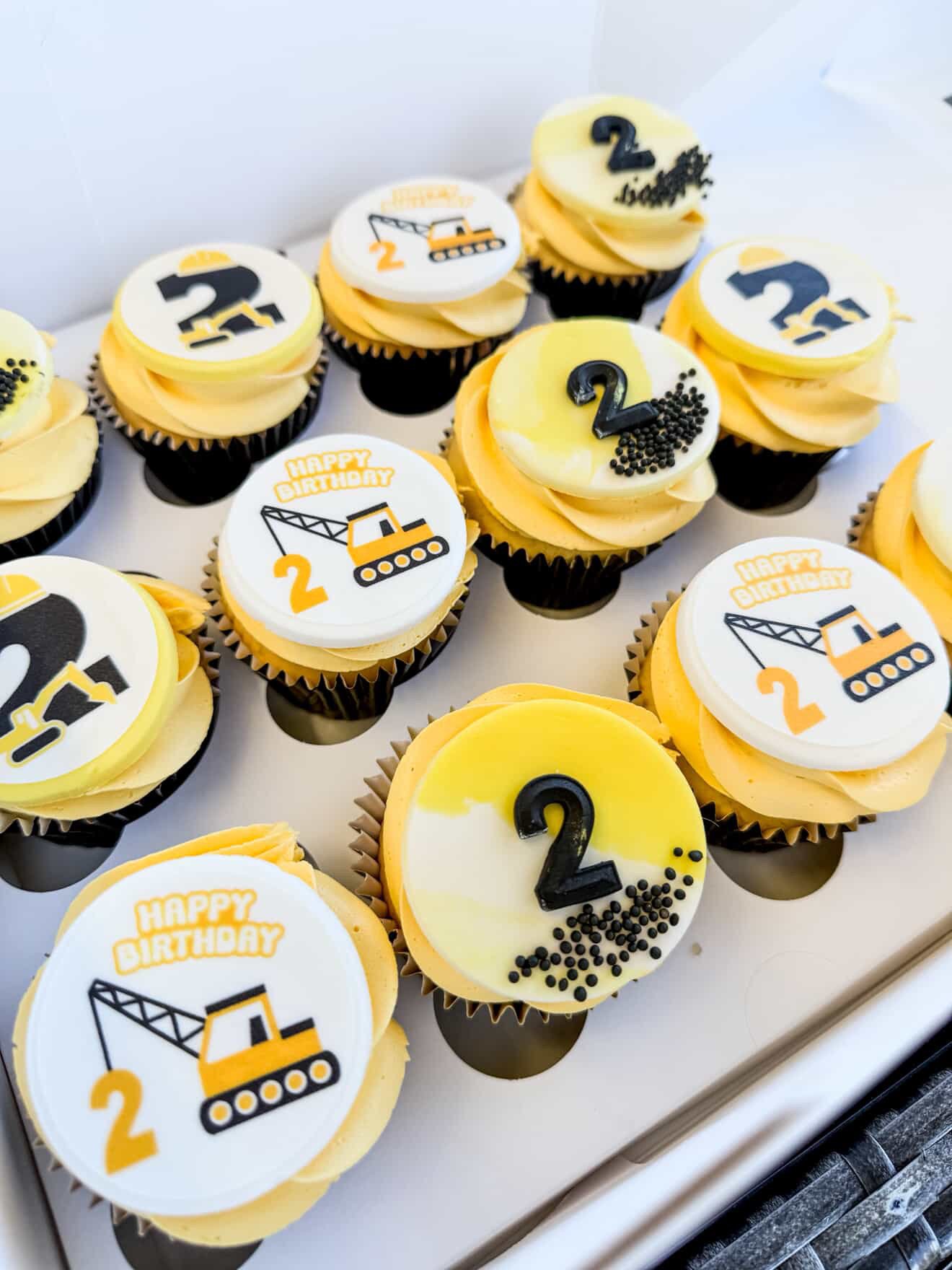 Cupcakes decorated with construction-themed toppers, featuring images of a crane and a dump truck, with yellow and black construction colors, and some cupcakes with black sprinkles.