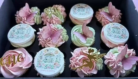 Cupcakes decorated with pink and green frosting, some with unicorn toppers and one with a "love" topper, in a black box.