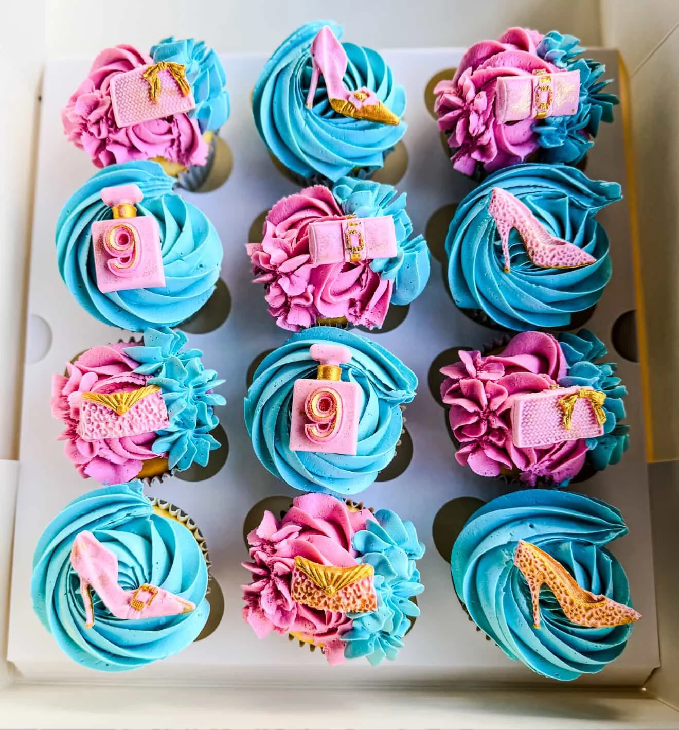 A box of cupcakes decorated with colorful pink and blue frosting and themed toppers, including high heels, champagne bottles, and the number 9, celebrating a birthday or special occasion.