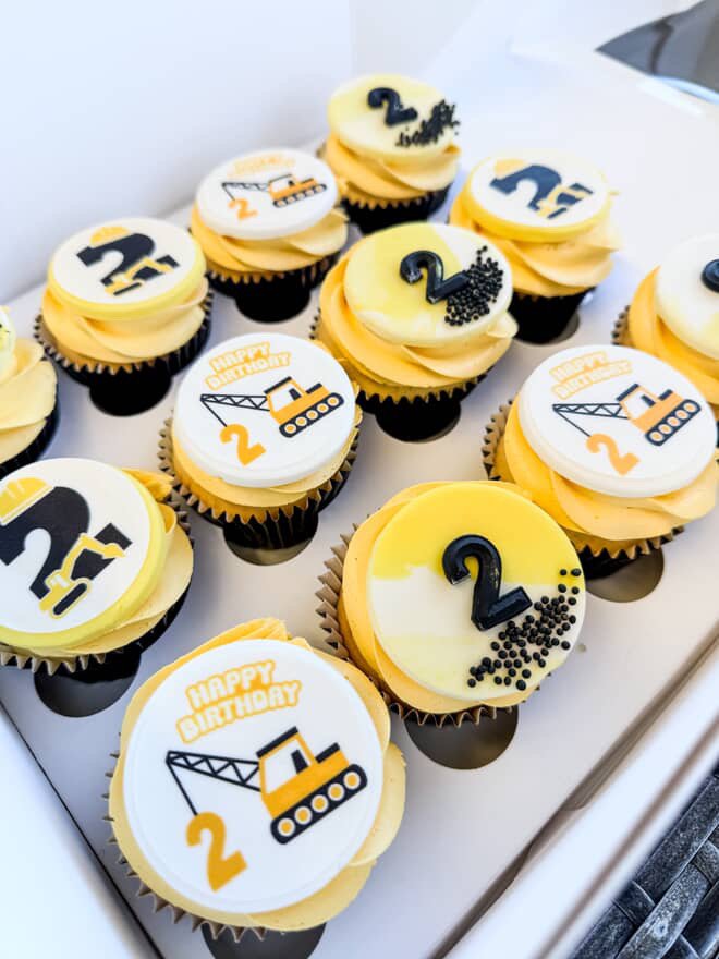 Cupcakes with construction vehicle theme decorations, including black and yellow color accents and number toppers, celebrating a second birthday.