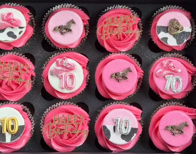 Pink cupcakes with various birthday-themed decorations, including candles, numbers, and animal toppers, arranged in a tray.