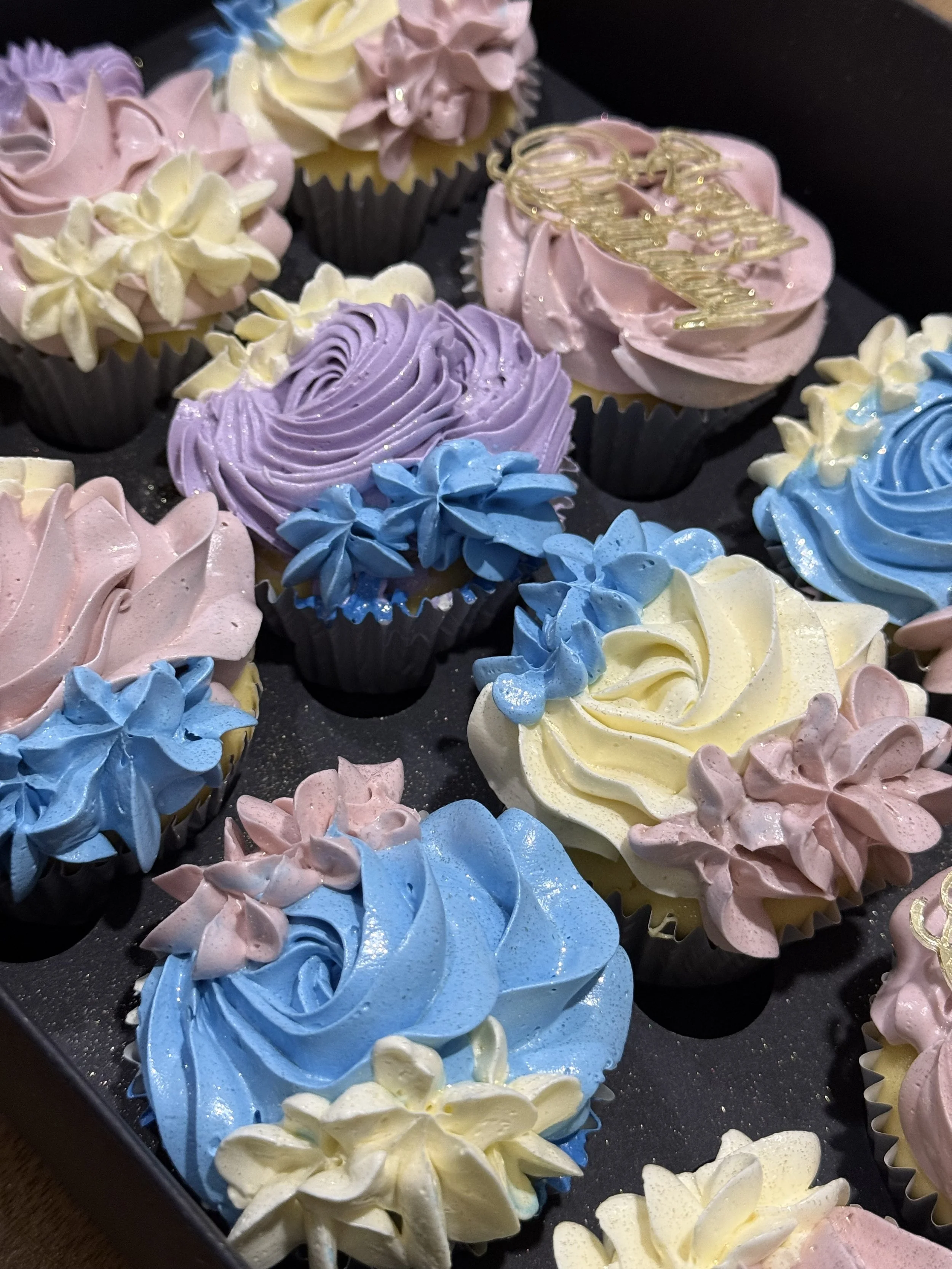 Assorted colorful cupcakes decorated with swirls of pastel pink, yellow, blue, purple, and white frosting, and some with floral decorations or edible gold accents, arranged on a black tray.