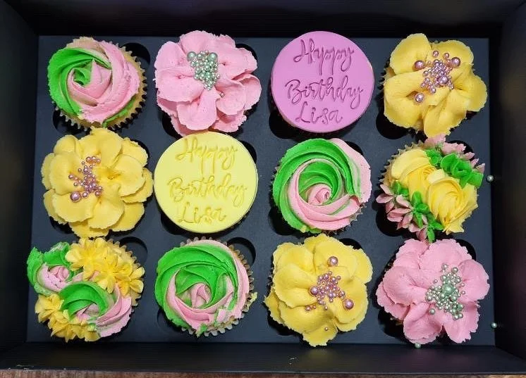 A box of decorated cupcakes with yellow, pink, and green frosting, decorated with pearl and sugar bead sprinkles, including two cupcakes with birthday messages for Lisa.