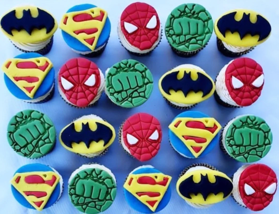 Cupcakes decorated with superhero logos: Batman, Superman, Spider-Man, Incredible Hulk, with cupcakes arranged in a grid.
