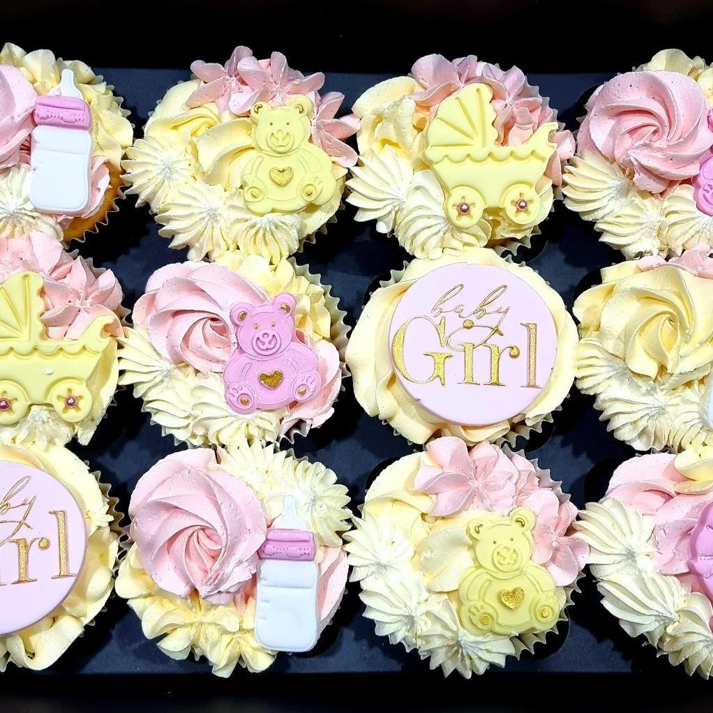 Assorted cupcakes decorated with pink and yellow frosting, some with teddy bear and baby carriage toppers, and one with the words "baby girl" written in gold on a pink disc.