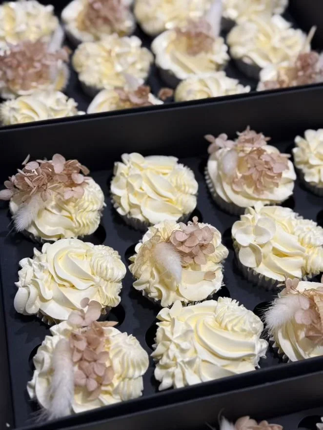 Tray of cupcakes decorated with white frosting, beige flower toppings, and small feathers.