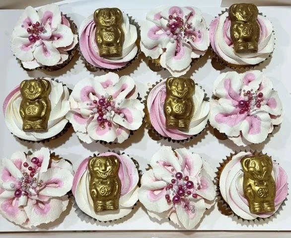 Decorative cupcakes with pink and white frosting, gold bear-shaped candies, and pink pearl sprinkles arranged in a grid pattern.