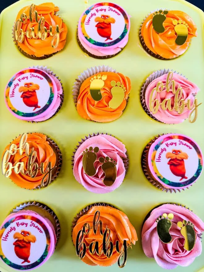 Collection of twelve cupcakes with orange or pink frosting, decorated with either 'oh baby' or 'Hunny Bunny' toppers, some featuring gold baby footprints or booties.