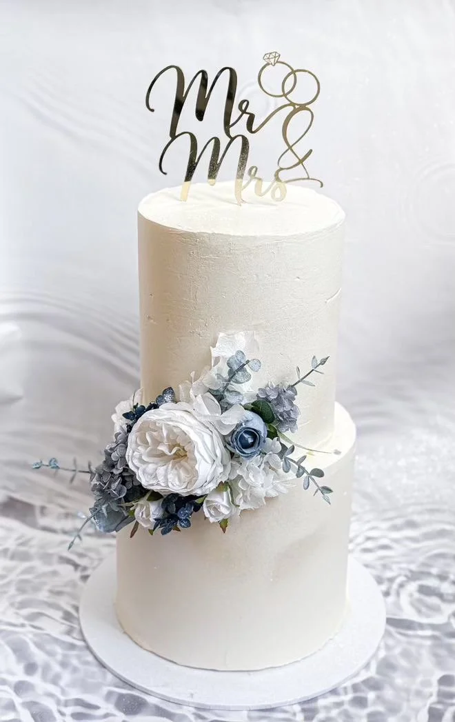 Two-tier white wedding cake with white and blue flowers and a topper with rings and 'Mr & Mrs' written in gold.