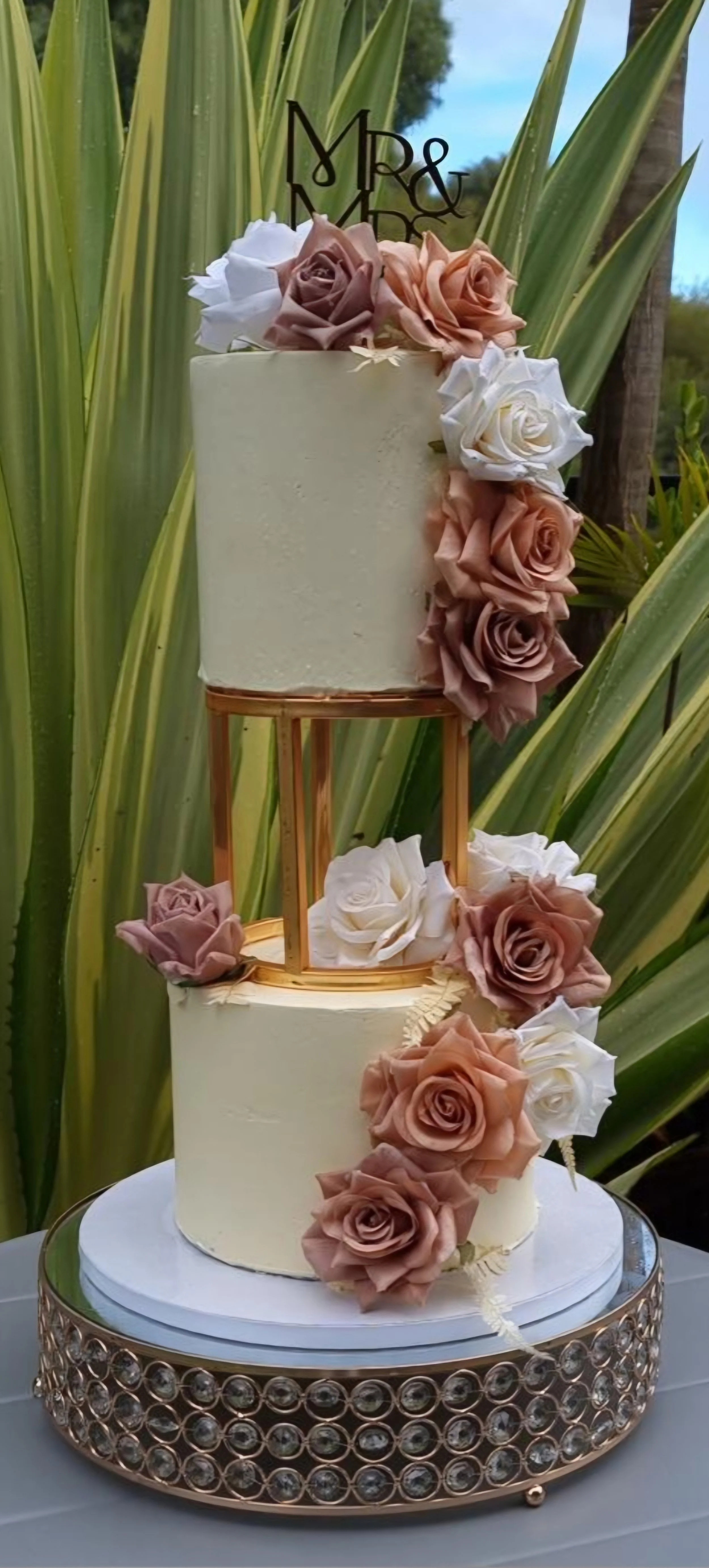 Two-tier white wedding cake decorated with pink and white roses, displayed outdoors with large green leaves in the background.