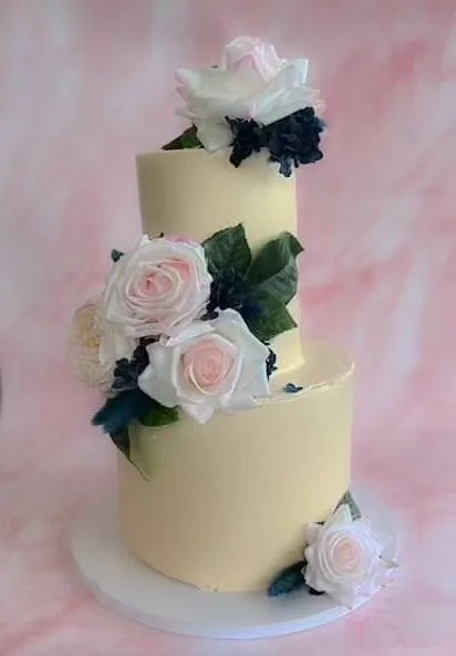 Two-tiered white wedding cake decorated with pink and white roses and dark greenery, against a soft pink background.