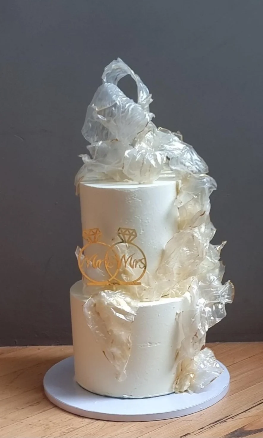 Three-tiered wedding cake decorated with gold accents, topped with white decorative elements resembling pearls or ruffled fabric.