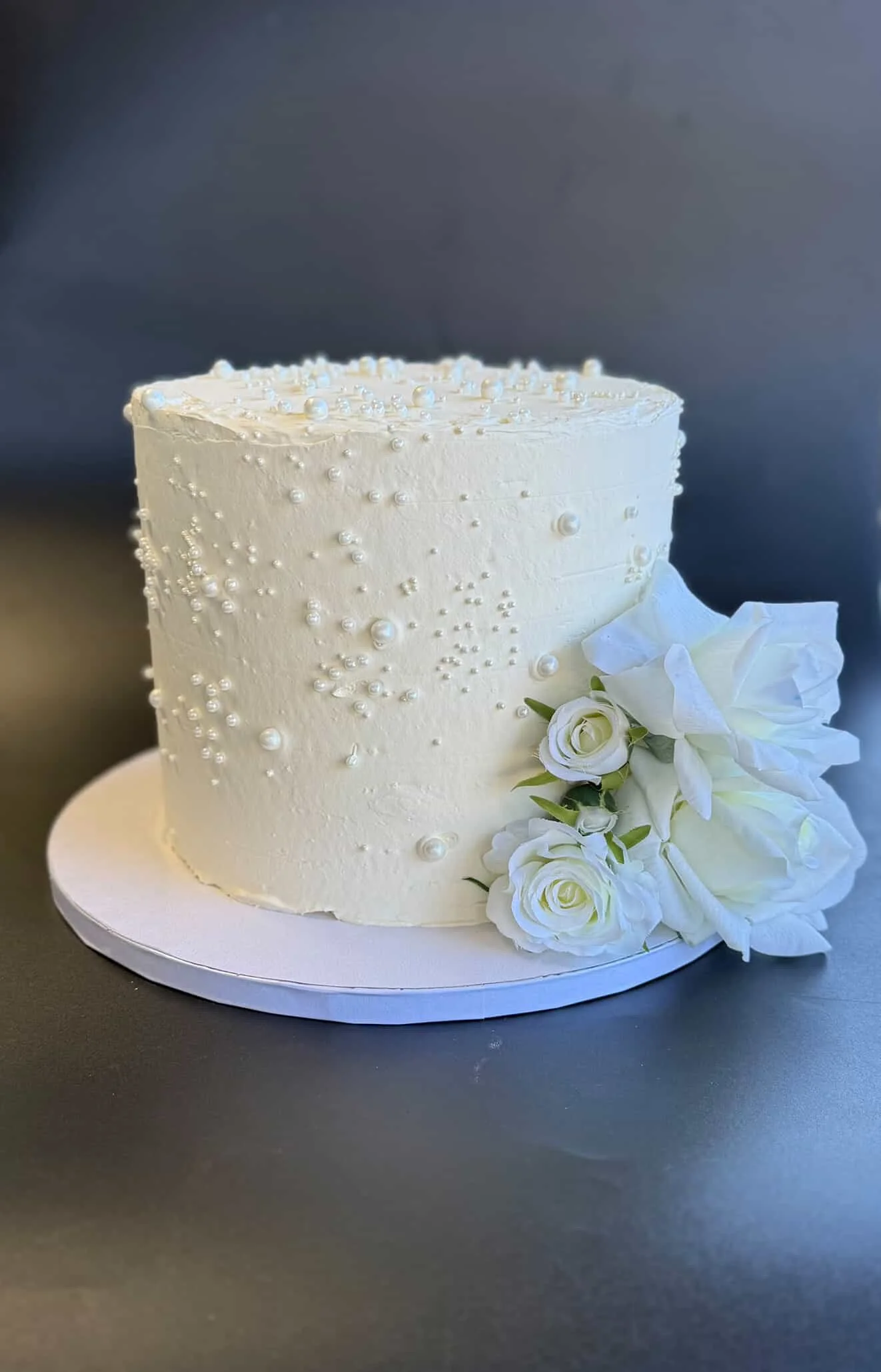 A white wedding cake decorated with pearls and white flowers, placed on a white cake board on a black surface with a dark background.