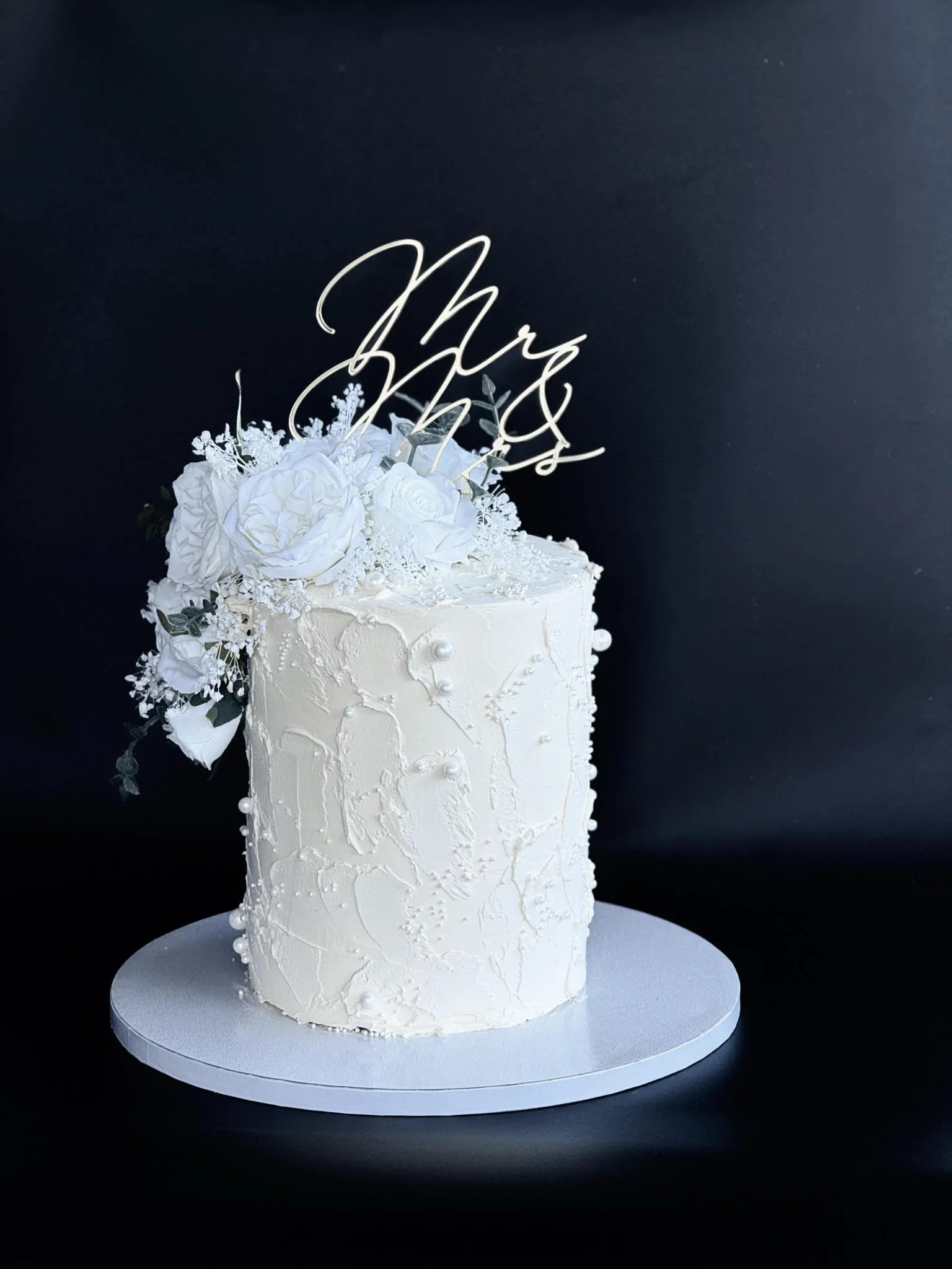 White wedding cake with floral decorations and a metallic cake topper that appears to spell out 'Mr & Mrs' on a silver cake board against a dark background.