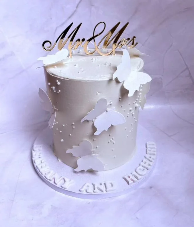 A white wedding cake with butterfly decorations, topped with a "Mr & Mrs" cake topper, placed on a white round base with the inscription "HAPPILY AND EVER AFTER".