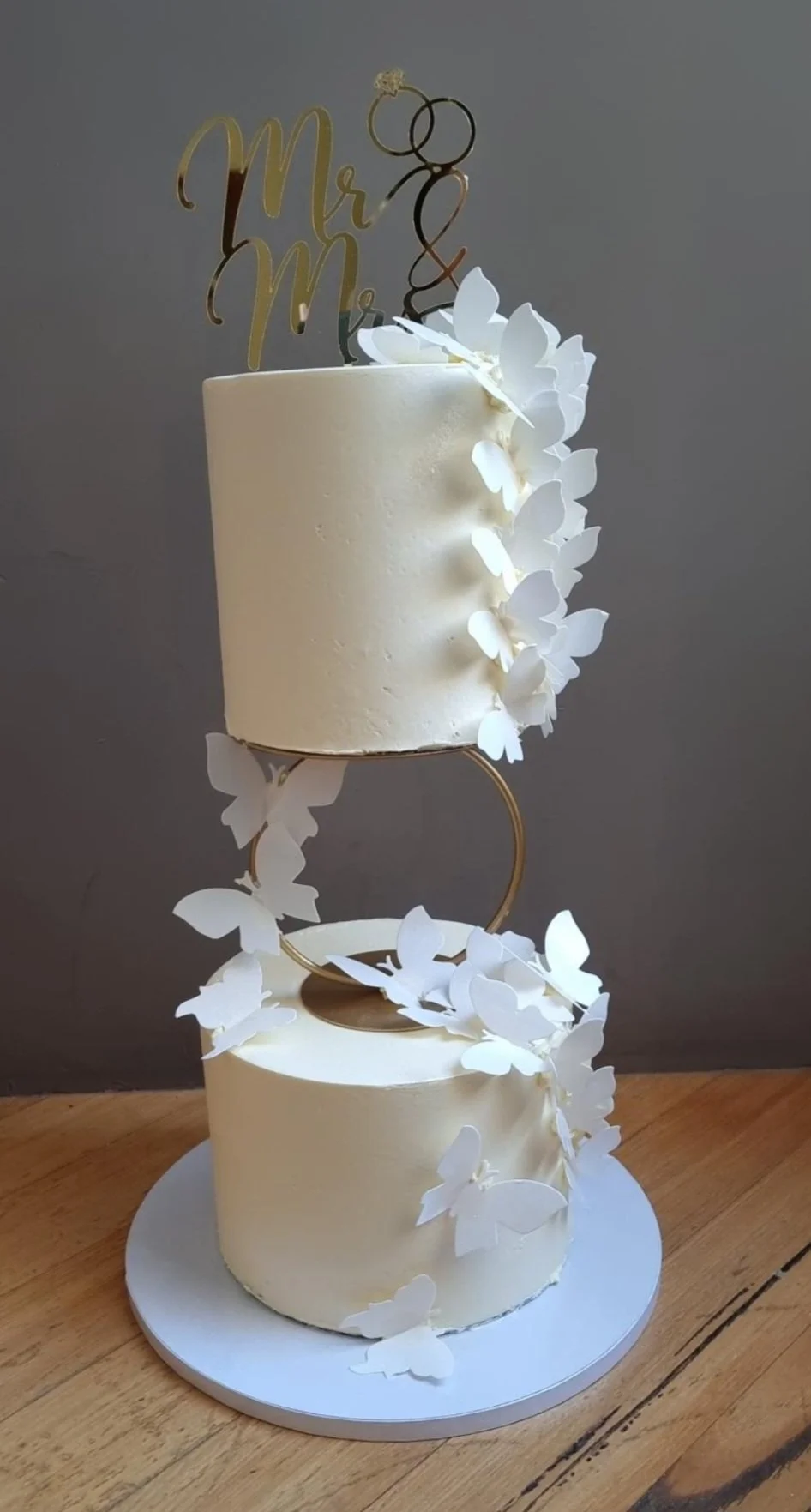 Two-tiered wedding cake with white buttercream frosting, decorated with white paper butterflies cascading from the top to the base, and a gold 'Mr & Mrs' cake topper.