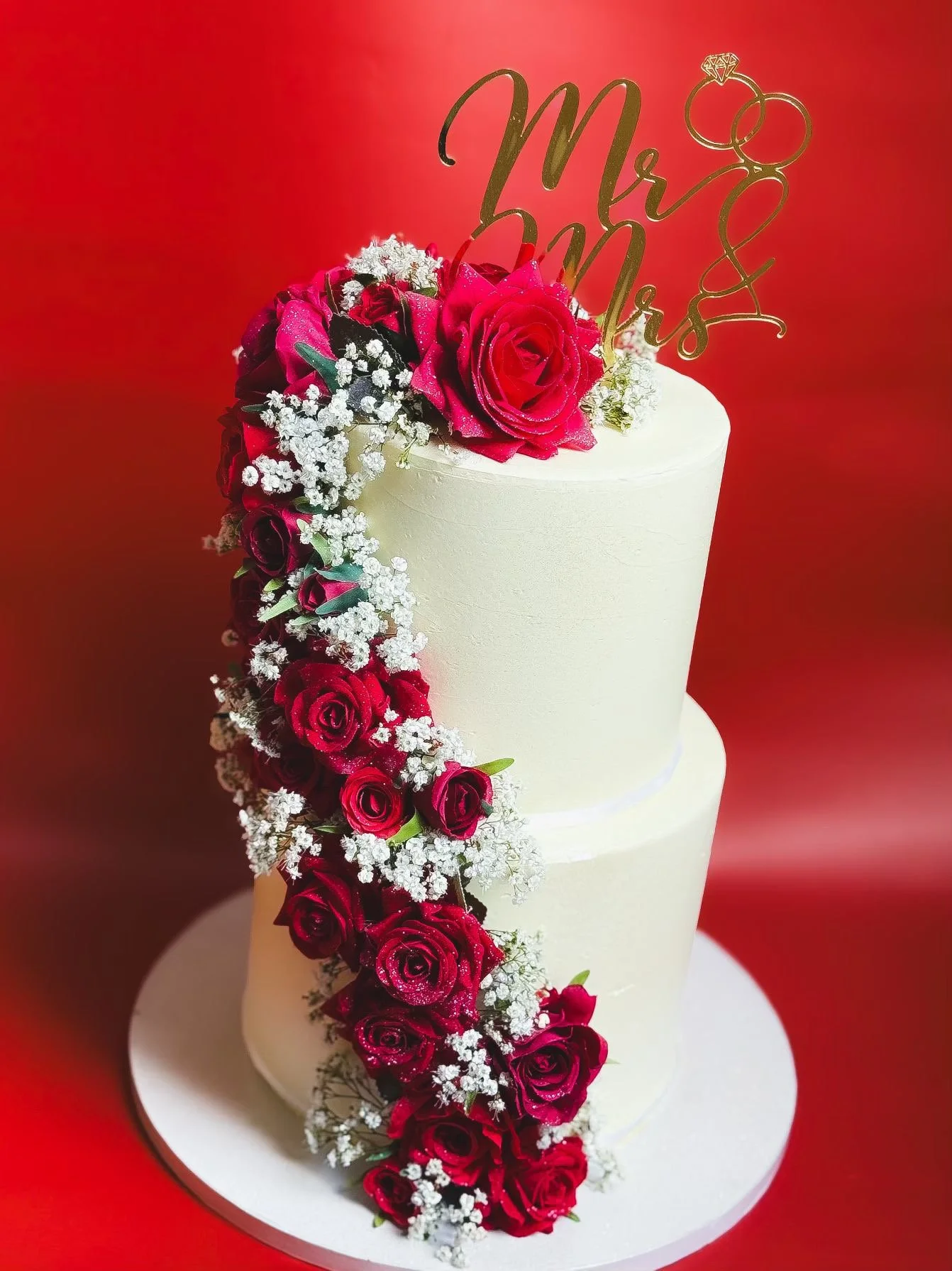 White wedding cake adorned with cascading red roses and white baby's breath, with a gold 'Mr & Mrs' cake topper with rings on a red background.