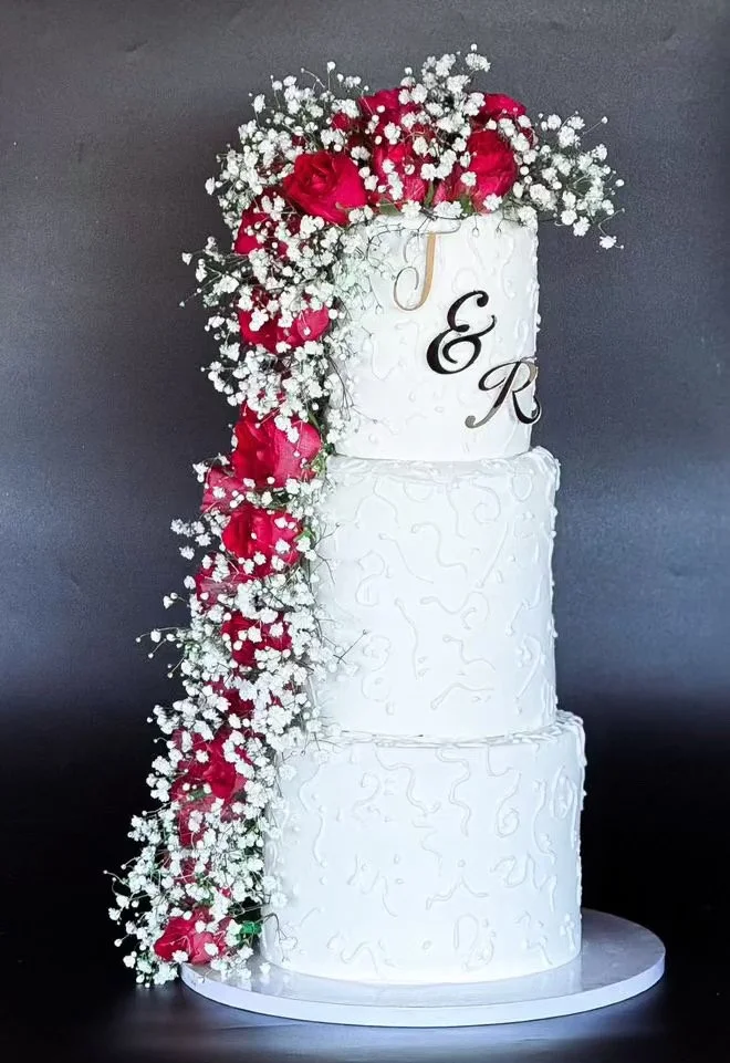 Three-tier white wedding cake decorated with white piping and floral arrangements of red roses and white baby's breath cascading from the top to the side.