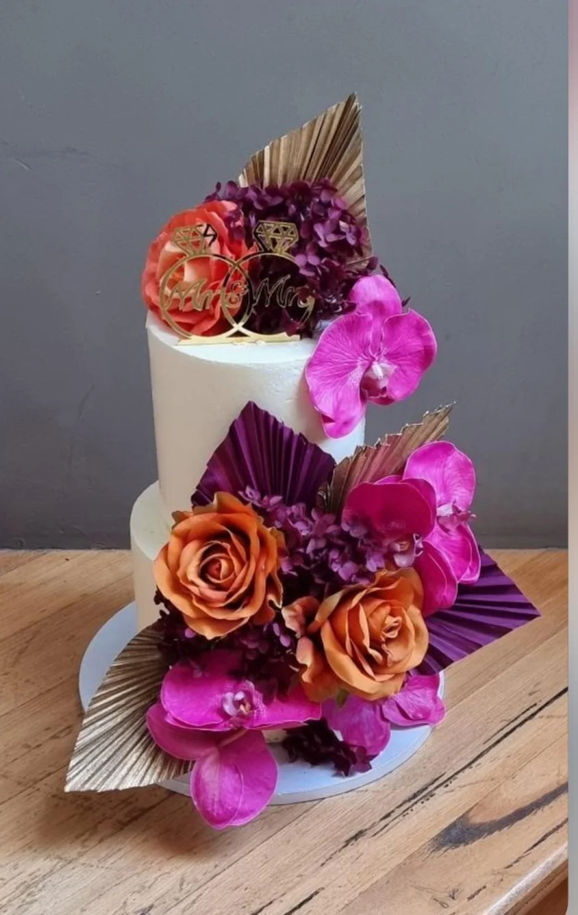 A two-tiered white wedding cake decorated with orange roses, pink orchids, purple and gold leaves, and gold wedding topper with rings and "Mr & Mrs" text.