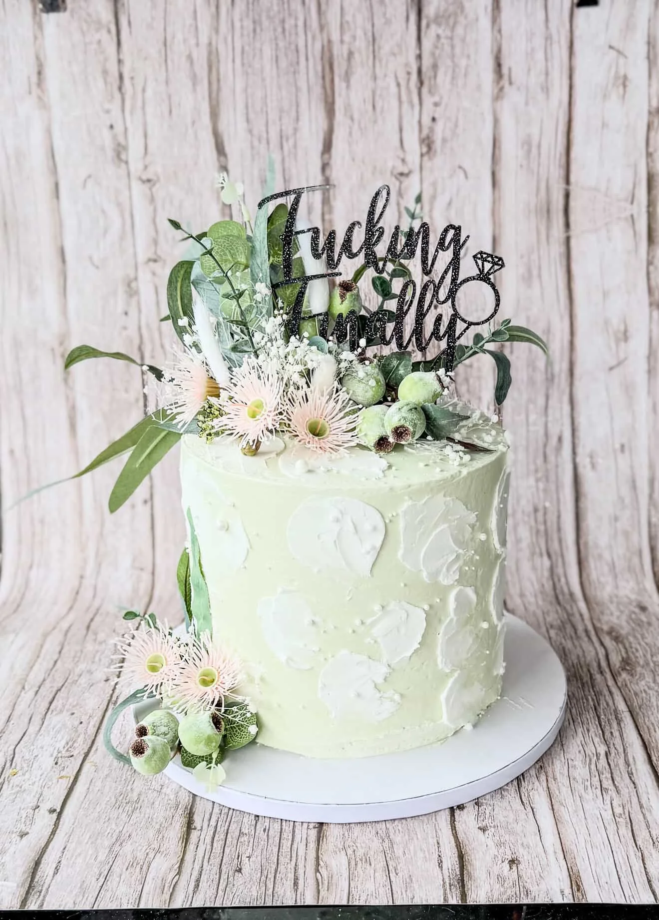 A decorated wedding or celebration cake with white icing, floral decoration, and a topper that says 'Fucking Tired' with a ring icon and diamond illustration, set on a wooden surface with a rustic wooden wall background.