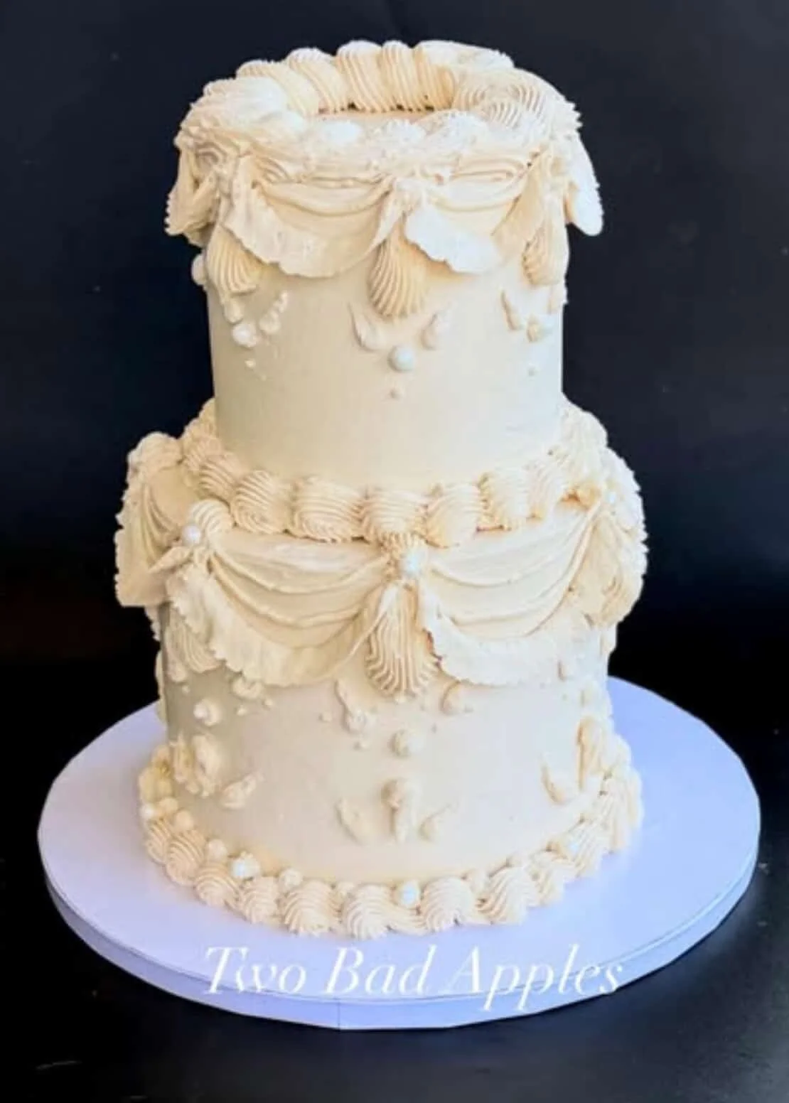 Three-tier white wedding cake decorated with piped frosting swags, dots, and rosette borders, displayed on a white cake board.