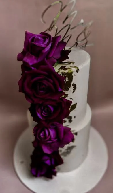 A white two-tiered cake decorated with dark purple roses and gold accents, topped with a spiral gold cake topper.