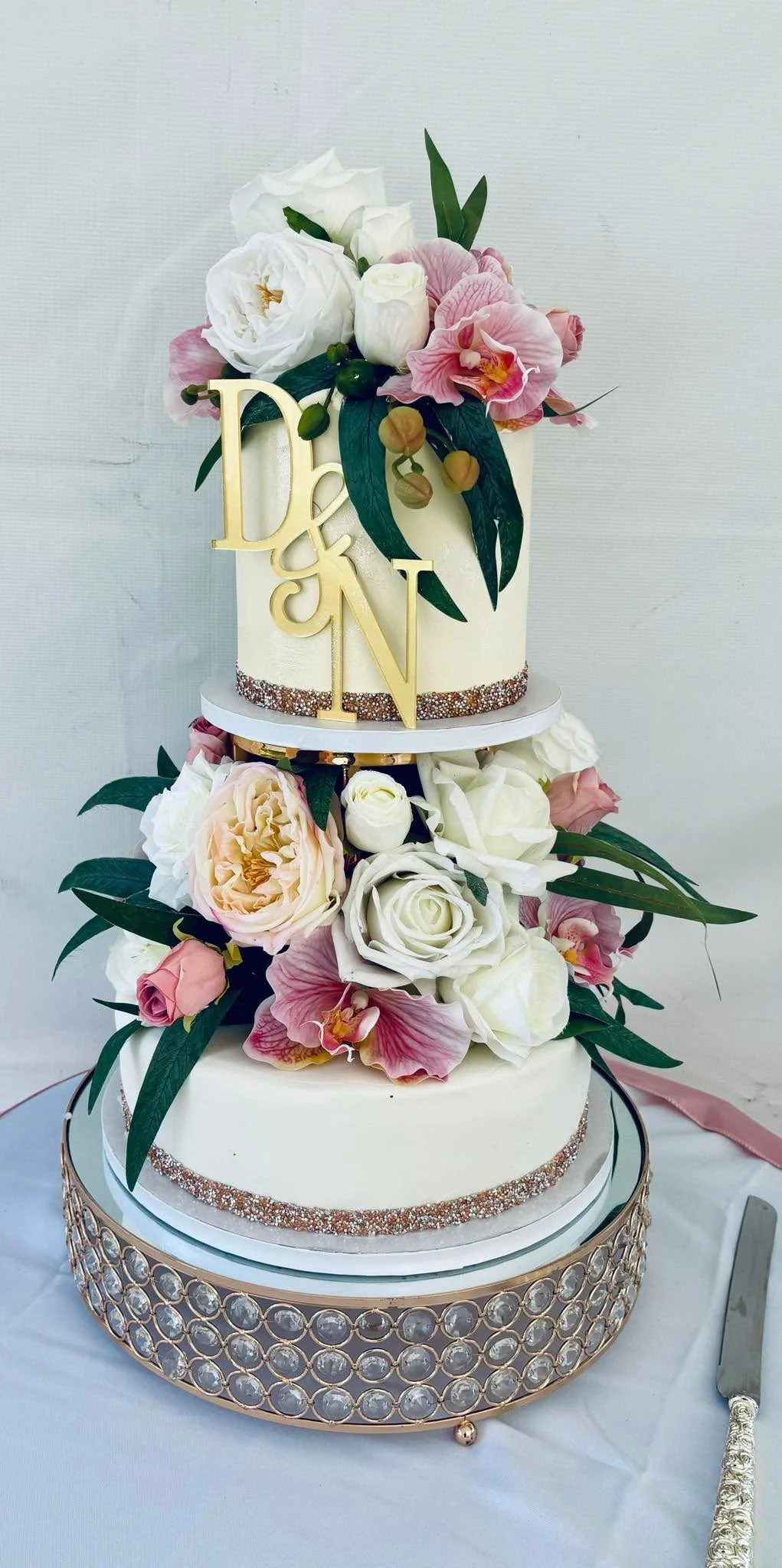 A two-tier wedding cake decorated with white and pink flowers, greenery, and a gold "Mrs. & Mrs." cake topper, set on a decorative stand with rhinestones.