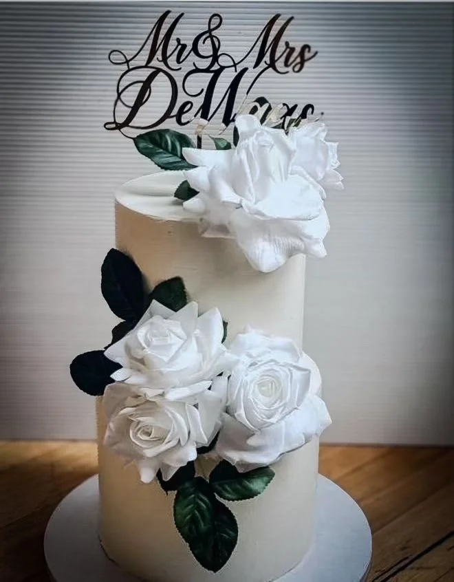A wedding cake decorated with white roses and green leaves, topped with a black 'Mrs & Mrs DeWarde' sign, placed on a round silver cake board.
