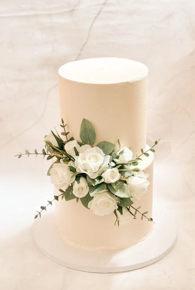 A white, single-tier wedding cake decorated with a cluster of white roses and green foliage around the middle, placed on a white round base against a light-colored marble background.