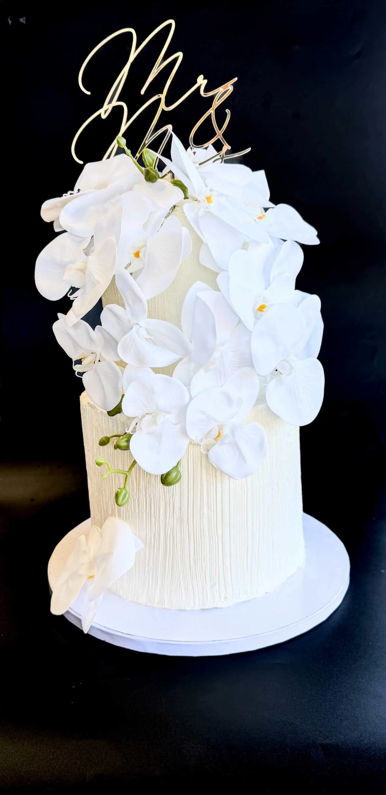 White cake decorated with cascading white orchids and a gold "Mrs" cake topper on a black background.