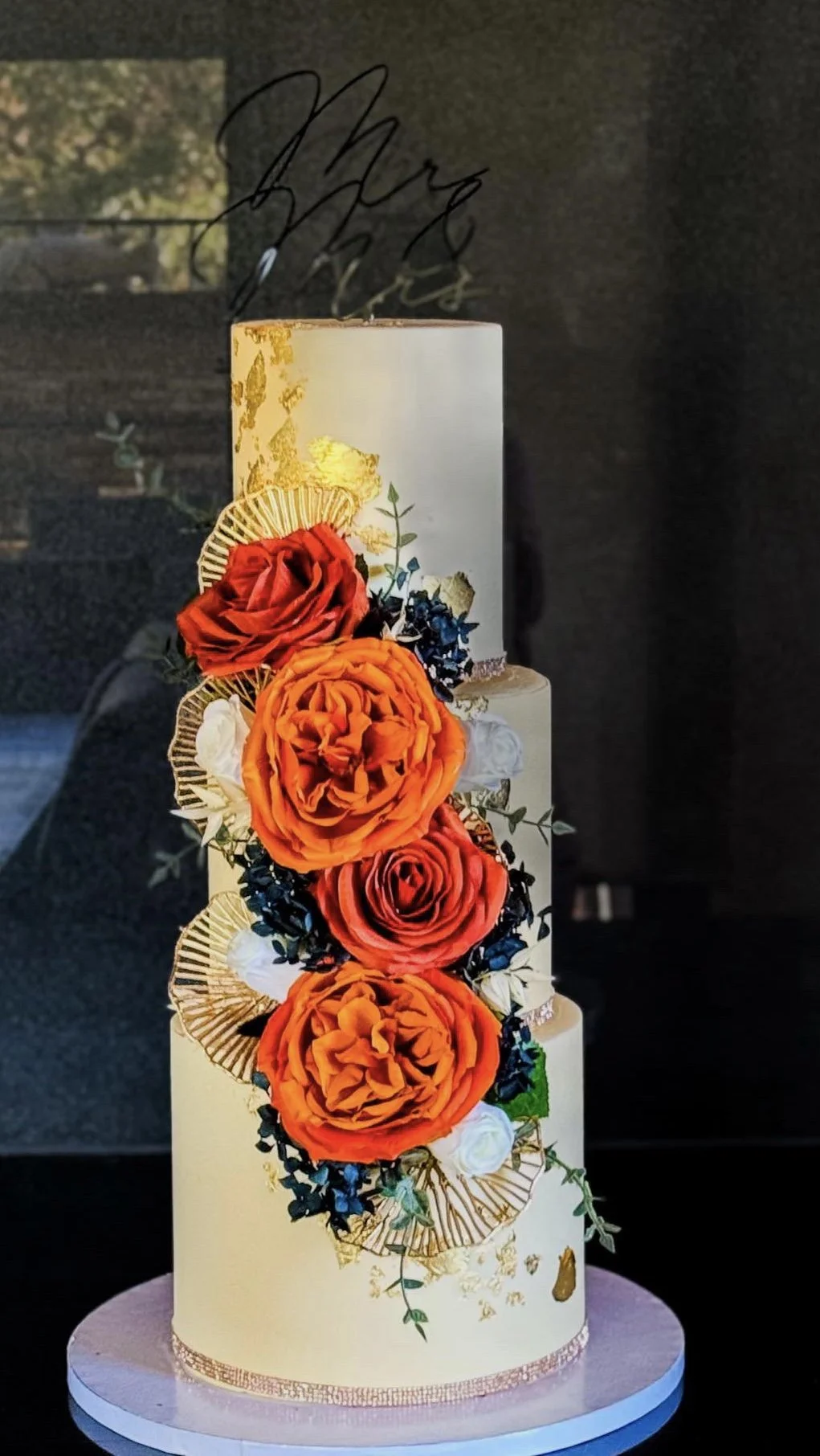 A tall three-tiered wedding cake decorated with large orange and red roses, white flowers, and black accents, with gold decorative elements and a black cursive cake topper.